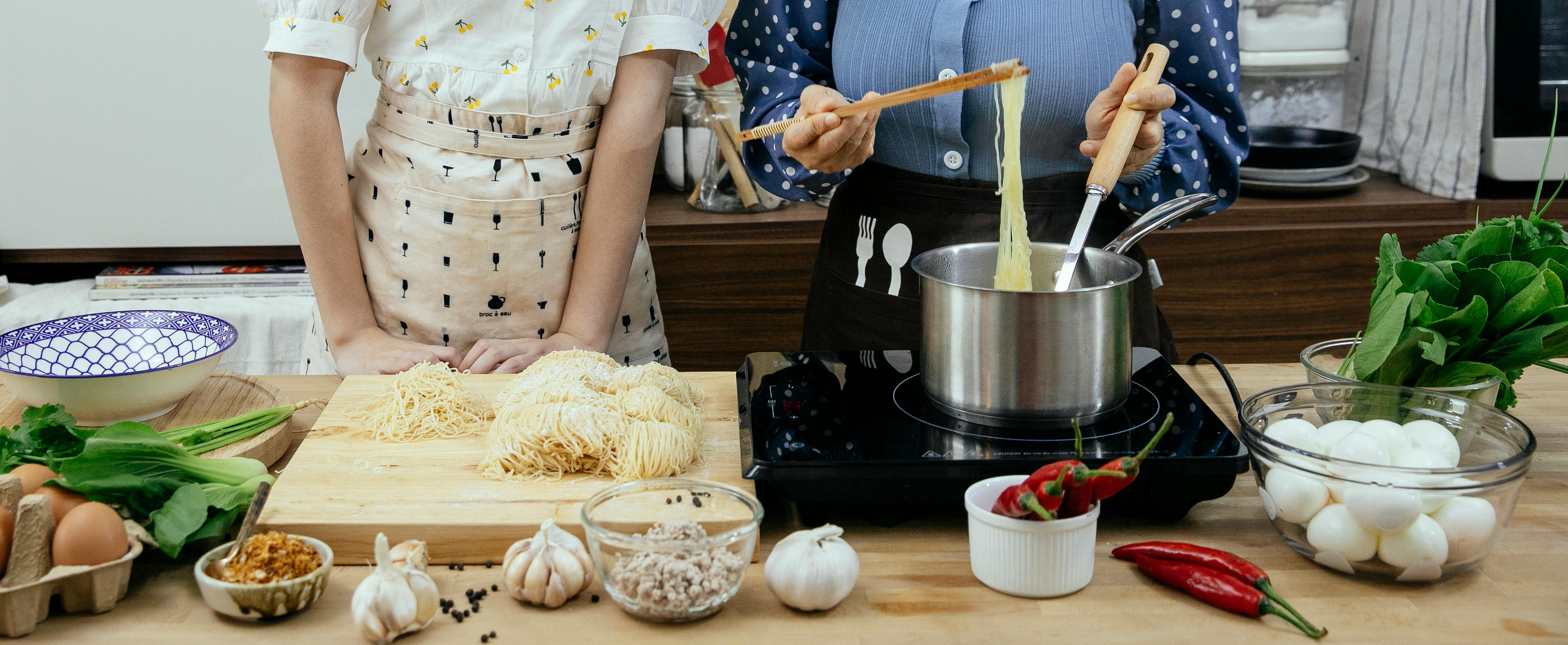 Unrecognizable female cooks with chopsticks cooking noddles in pan on stove while standing at table with raw pasta on cutting board
